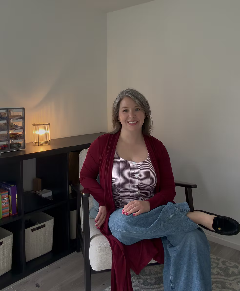 A woman with shoulder-length gray hair, wearing a red cardigan and striped top, sitting on a chair in a room with a beige wall, a black bookshelf, and a lamp in the background.