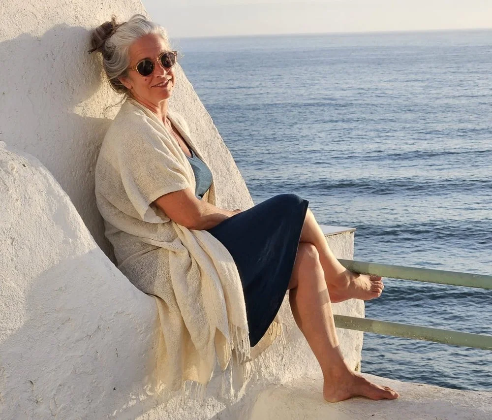 Eine Frau mit Sonnenbrille und in einem blauen Kleid sitzt entspannt an einer felsigen Küste mit Blick aufs Meer, umgeben von weißen Klippen.