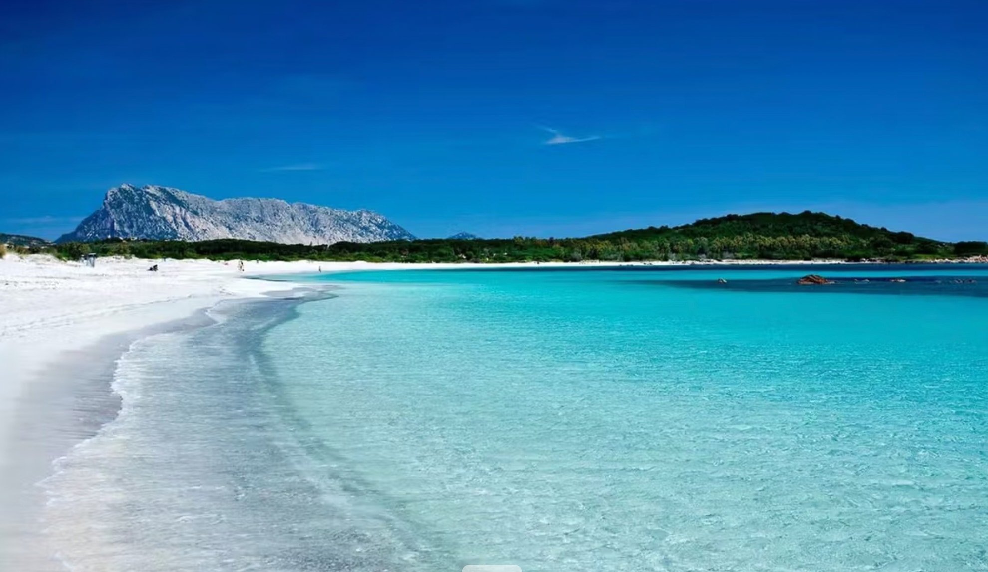 Blaue Meeresbucht mit weißem Strand, grünen Bäumen im Hintergrund und hohen Bergen bei klarem Himmel.