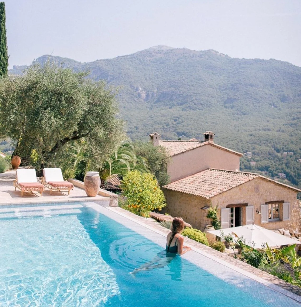 Frau entspannt im Pool mit Blick auf ein mediterranes Dorf mit Steinhäusern und grünen Bergen im Hintergrund.