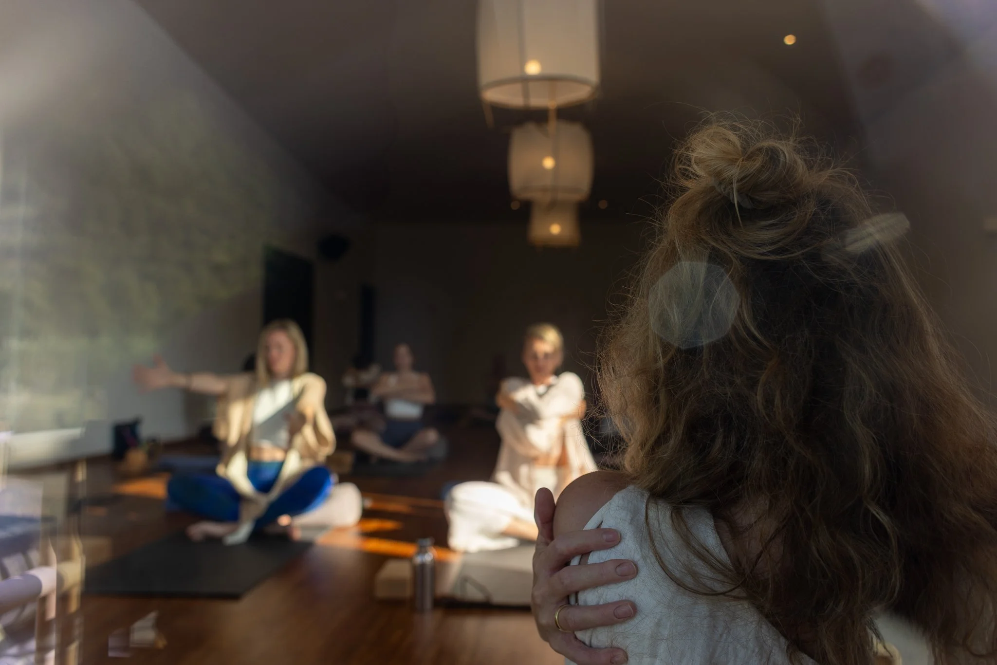 Frau mit wild durcheinander gewaschenen Haaren, die eine Umarmung mit einer anderen Person im Hintergrund hat, während eine Gruppe an Menschen in einem Raum sitzt, der für eine Yogastunde oder Meditation vorbereitet ist.