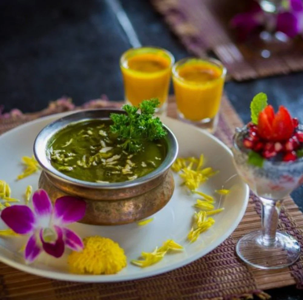 Ein indisches Mahl mit grünem Curry, zwei Orangensaft-Getränken, einem Dessert mit Beeren und einer Blume auf einem Teller, alles auf einem Holztablett serviert.