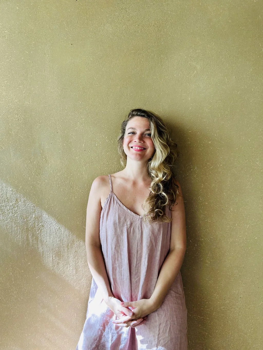 Frau mit lockigem Haar und einem rosa Kleid steht vor einer goldfarbenen Wand und lächelt.