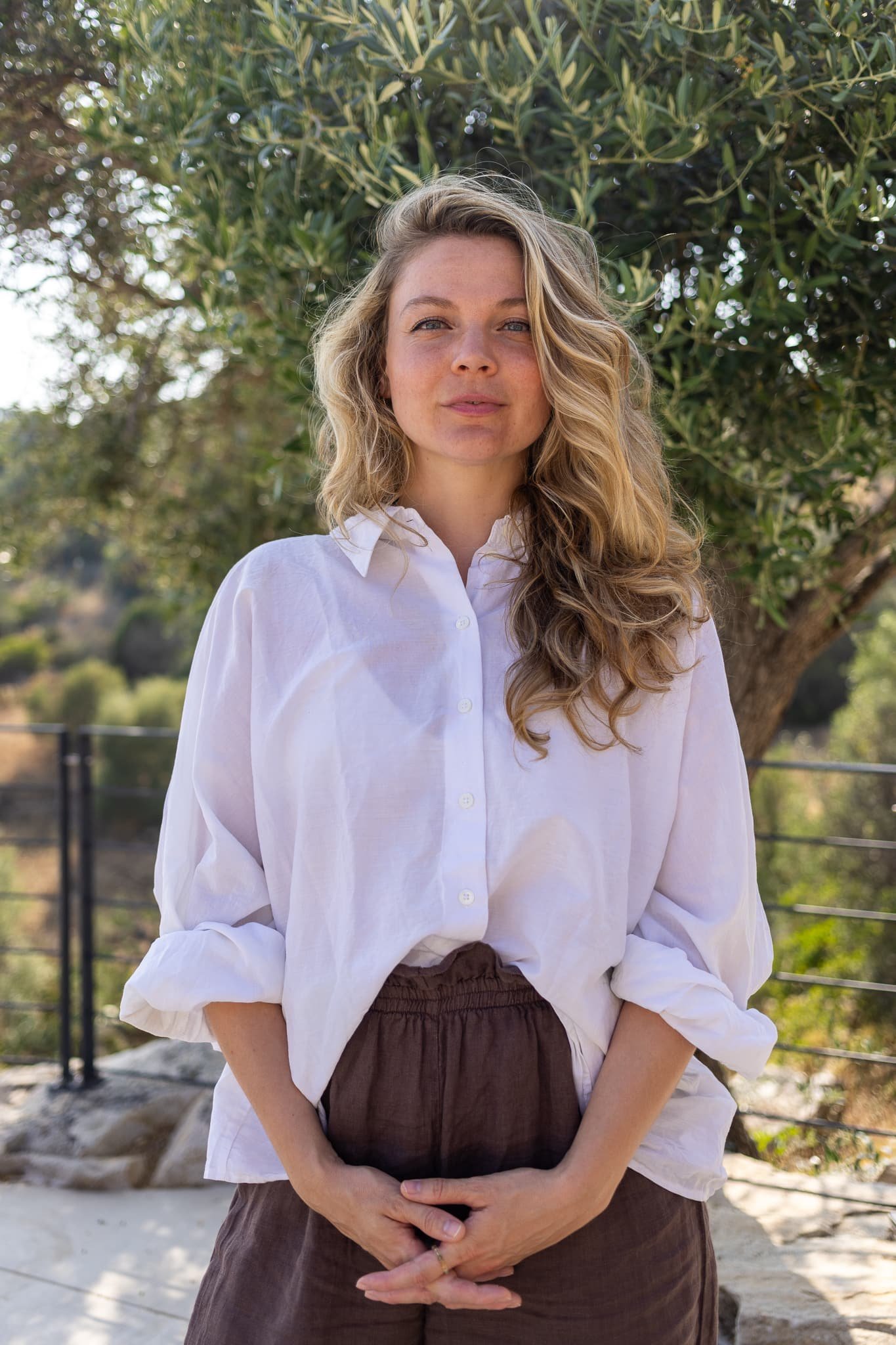 Eine junge Frau mit lockigem, blondem Haar trägt eine weiße Bluse und braune Bundfaltenhose, steht im Freien vor einem Baum. Sie hat einen neutralen Gesichtsausdruck.