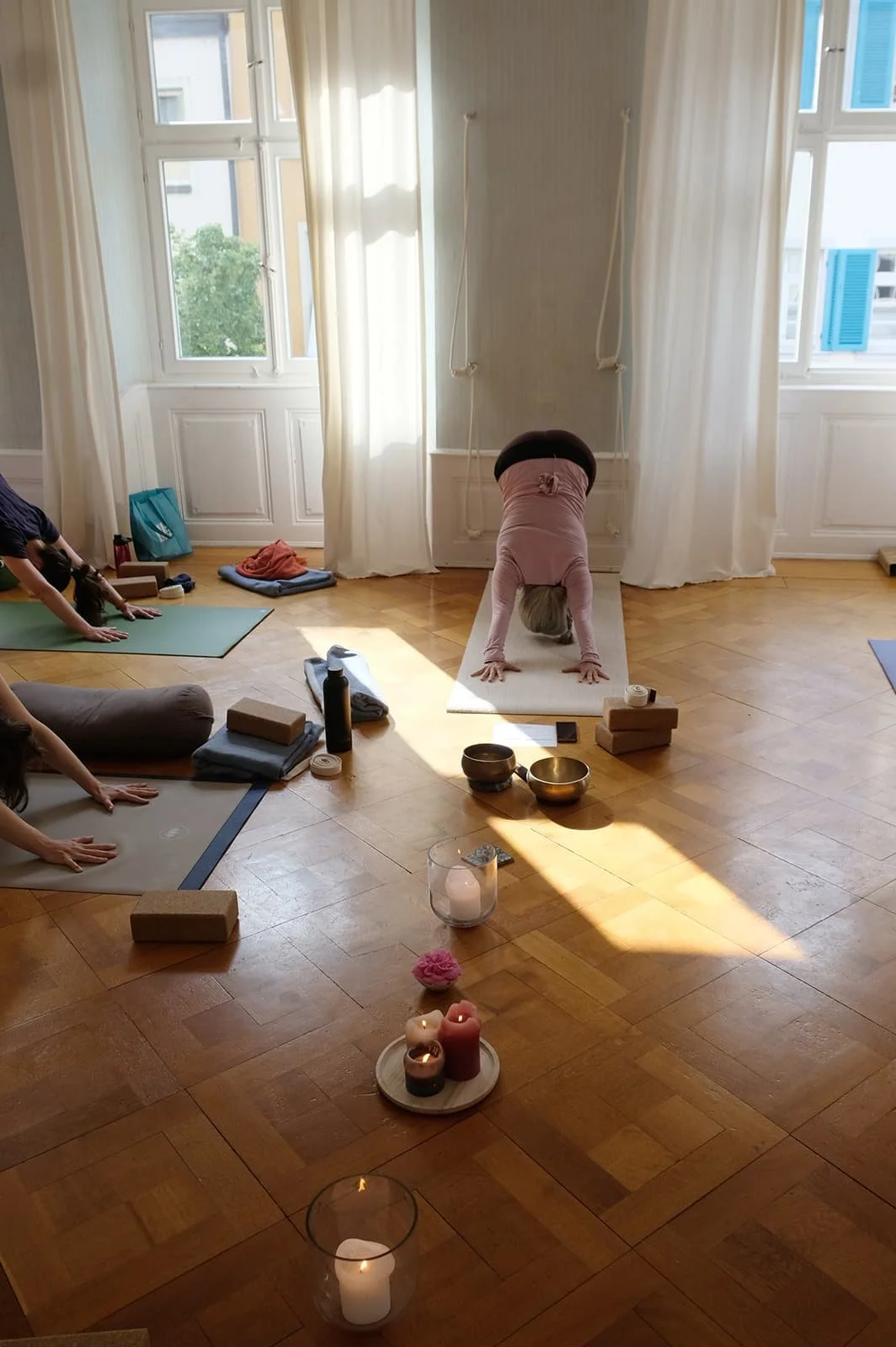 Menschen beim Yoga in einem hellen Raum mit großen Fenstern, Kerzen auf dem Boden, Yoga-Matten und Yogazubehör.