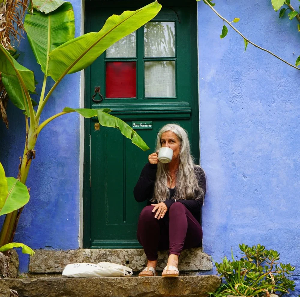 Eine Frau mit langen grauen Haaren sitzt vor einer blauen Hauswand auf einer Steintreppe und trinkt aus einer weißen Tasse. Es gibt große grüne Blätter im Vordergrund und einen grünen Türrahmen hinter ihr.