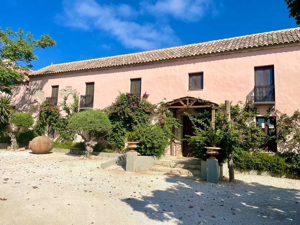 Ein rosa zweistöckiges Haus mit Ziegeldach, umliegend von Bäumen und Sträuchern, mit Steinstufen zum Eingang und einem klaren blauen Himmel im Hintergrund.