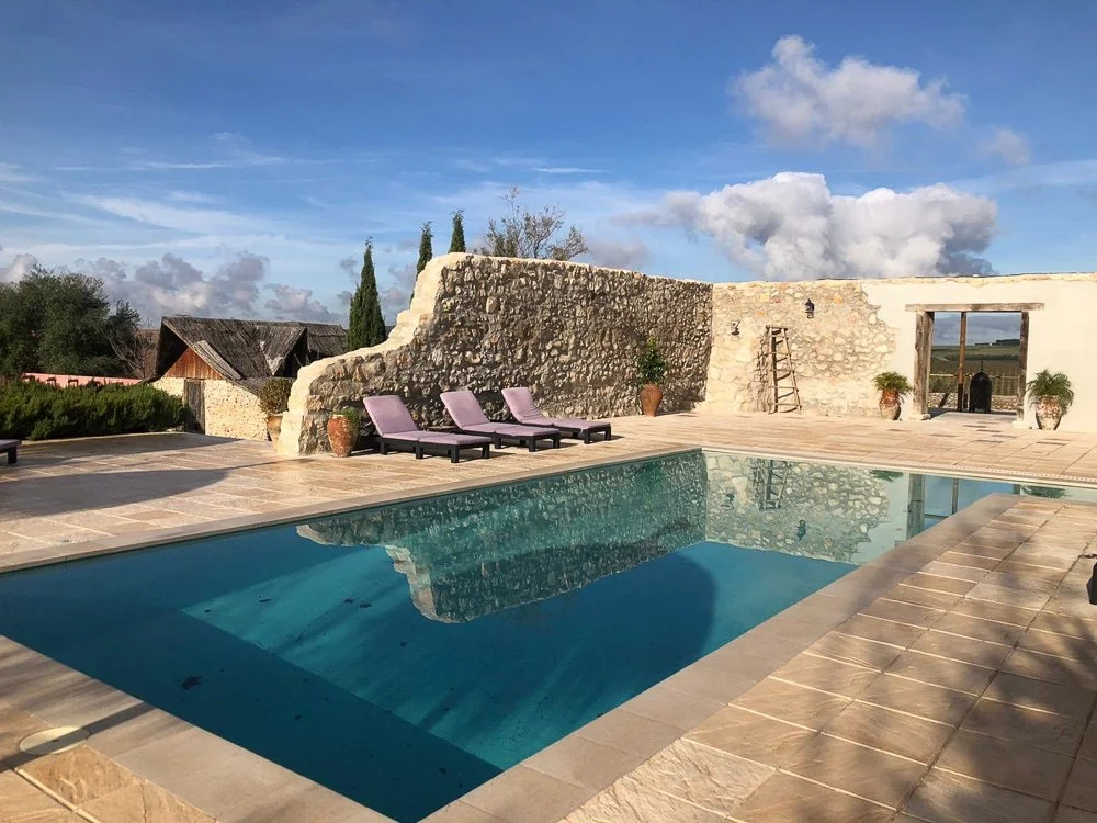 Schöne Poollandschaft mit vier Liegestühlen, einer alten Steinmauer, Töpfen mit Pflanzen und einem blauen Himmel mit Wolken im Hintergrund.