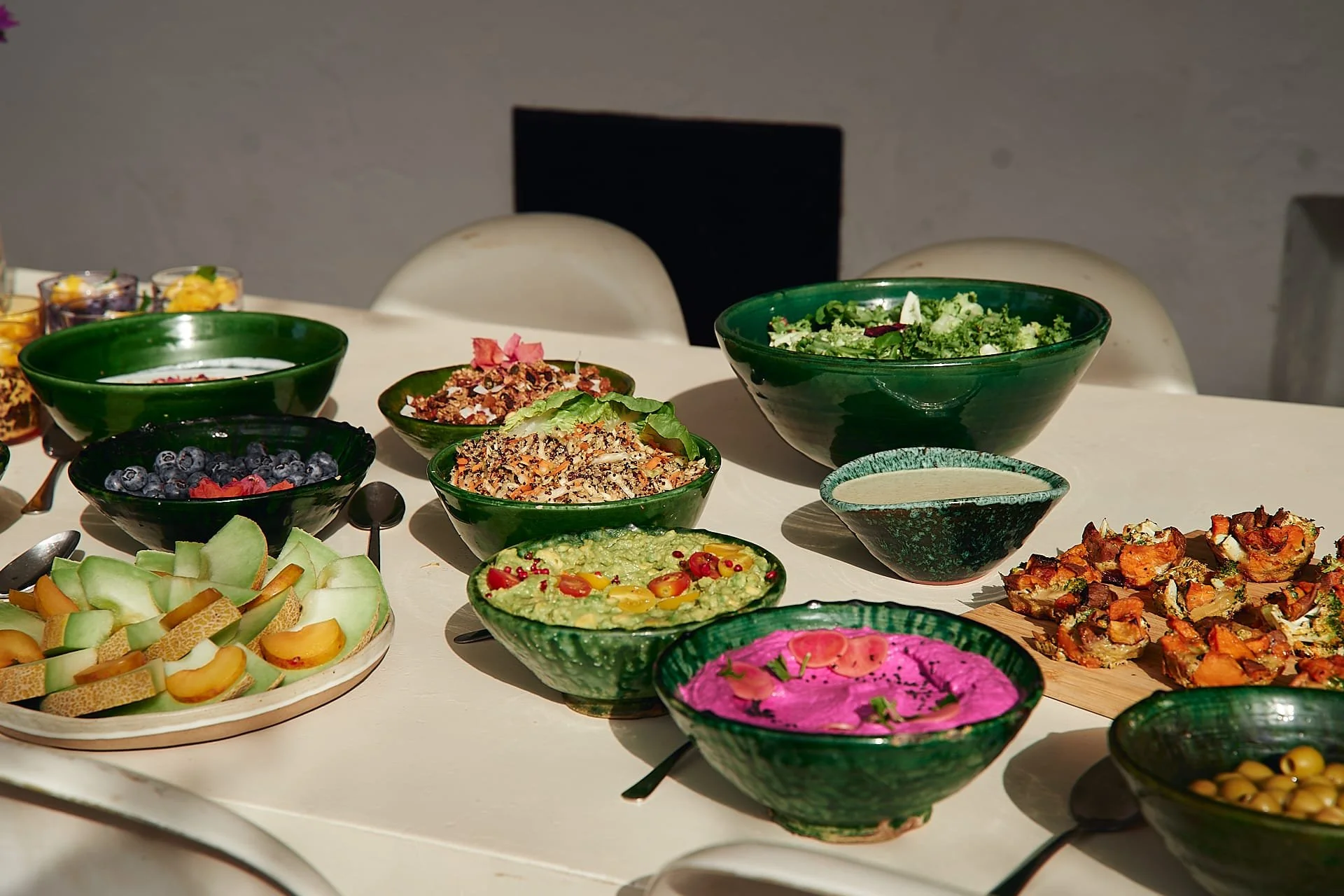 Verschiedene bunte Salate und Früchte in grünen Schüsseln auf einem weißen Tisch, vermutlich bei einem Buffet oder Festmahl.