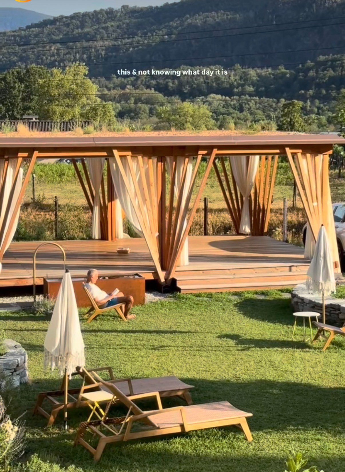 Ein Mann liest ein Buch in einem Garten mit Liegestühlen, einem Pavillon und grüner Natur im Hintergrund.