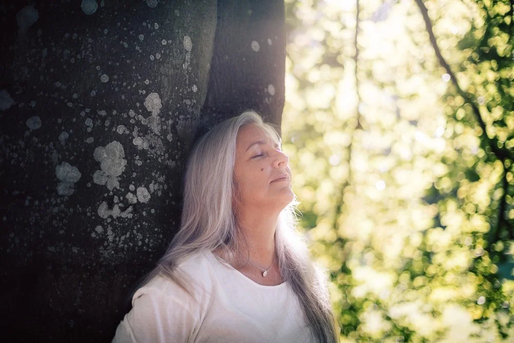 Frau mit langen grauen Haaren lehnt an einem Baum im Sonnenschein, entspannt und mit geschlossenen Augen.