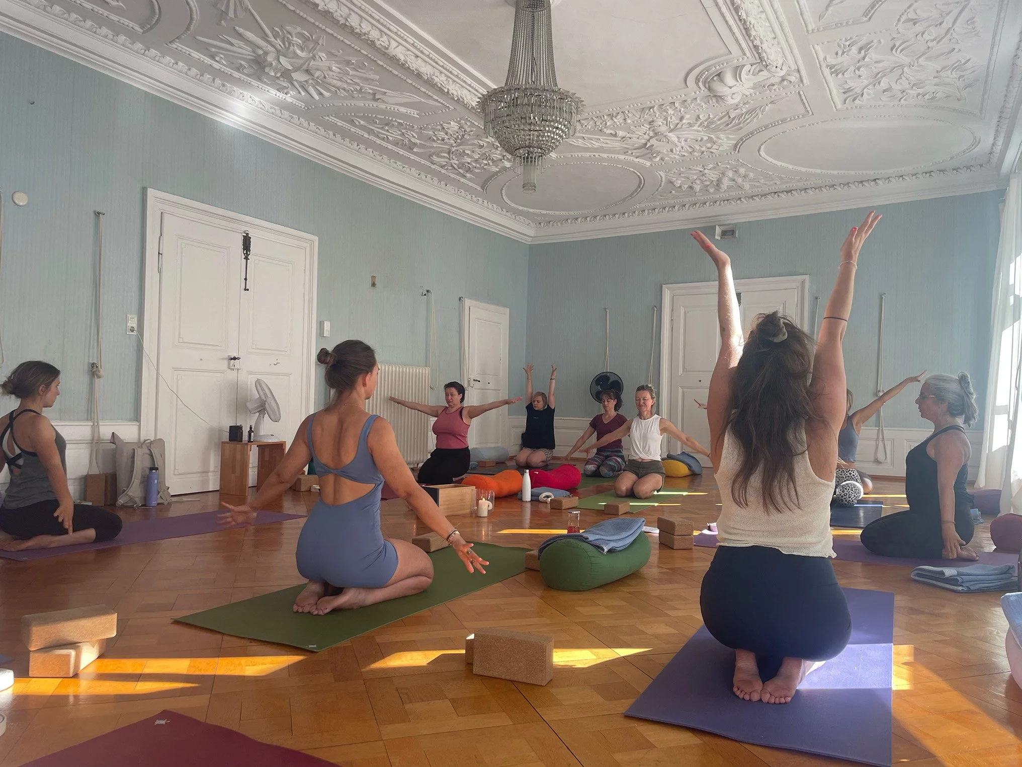 Menschen in einem Raum beim Yoga oder Meditation, sitzend auf Yogamatten in einer Gruppe. Es gibt diverse Yoga-Zubehör wie Kissen, Blöcke, Decken und Wasserflaschen. Der Raum hat eine hohe, verzierte Decke mit Kronleuchter und große Fenster, die natürliches Licht einlassen.