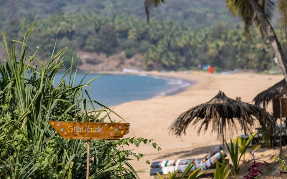 Strand mit Sonnenschirmen und Palmen, im Hintergrund Wasser und grüner Hügel, ein handgemachtes Holzschild mit der Aufschrift 'Gratitude'