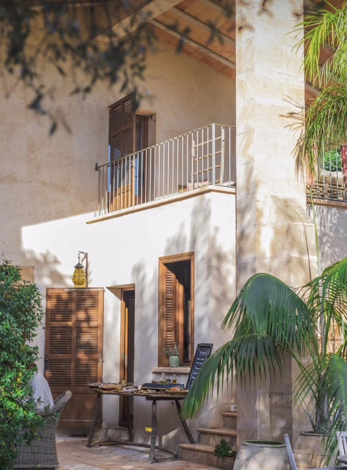 Ein Sommergarten mit Terrasse, Pflanzen und einem Gebäude im mediterranen Stil mit Holzfenstern und Balkonen. Es gibt einen Tisch mit Essen und einen Stuhl.
