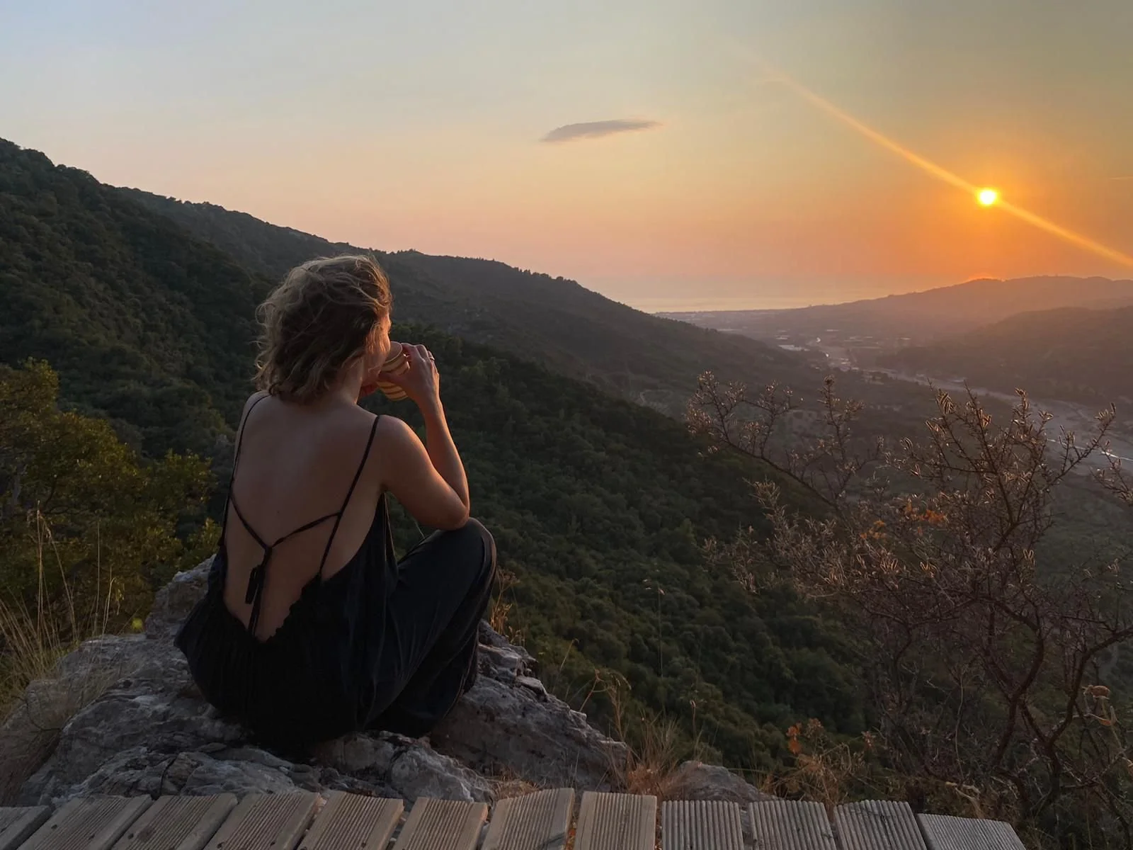 Eine Frau sitzt auf einem Felsen und trinkt aus einer Tasse, während sie den Sonnenuntergang über einer Hügellandschaft beobachtet.