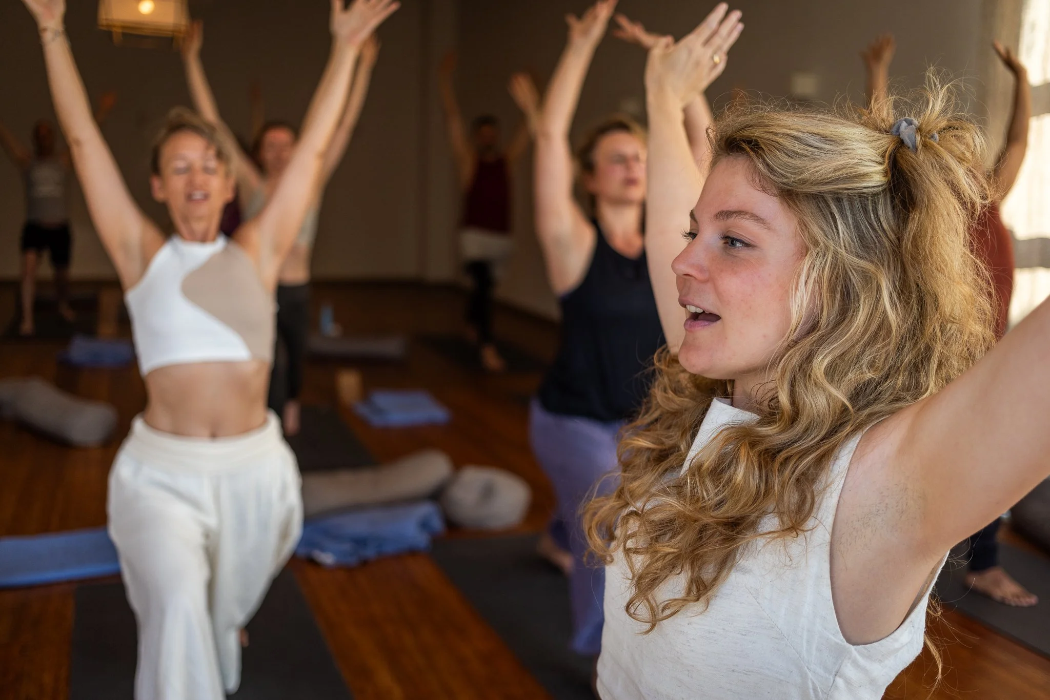 Junge Frau macht Yoga in einem Fitnessstudio, während im Hintergrund andere Frauen ebenfalls Yoga machen.