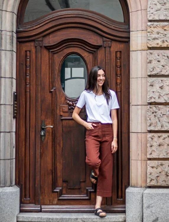 Junge Frau mit weißen T-Shirt und braunen Hosen, die vor einem großen, kunstvoll geschnitzten Holztor steht und lächelnd in die Kamera schaut.