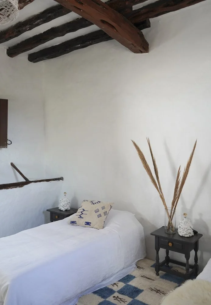Schlafzimmer mit weißem Bett, zwei dunkle Nachttische mit Dekorationen, Zierkissen, Holzdeckenbalken und getrockneten Gräsern.