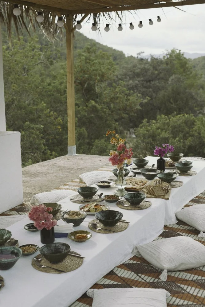 Ein Esstisch mit mehreren tellern, bowls und blumenvasen, auf einem Blick in der Natur, mit Sitzkissen auf Bodenniveau, unter einer offenen Holzdachkonstruktion mit Lichterketten.