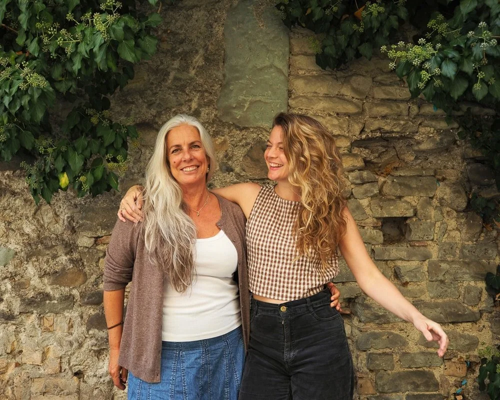 Zwei lachende Frauen, eine ältere mit langem grauem Haar und eine jüngere mit lockigem braunem Haar, umarmen sich vor einer Steinmauer mit grünen Pflanzen.