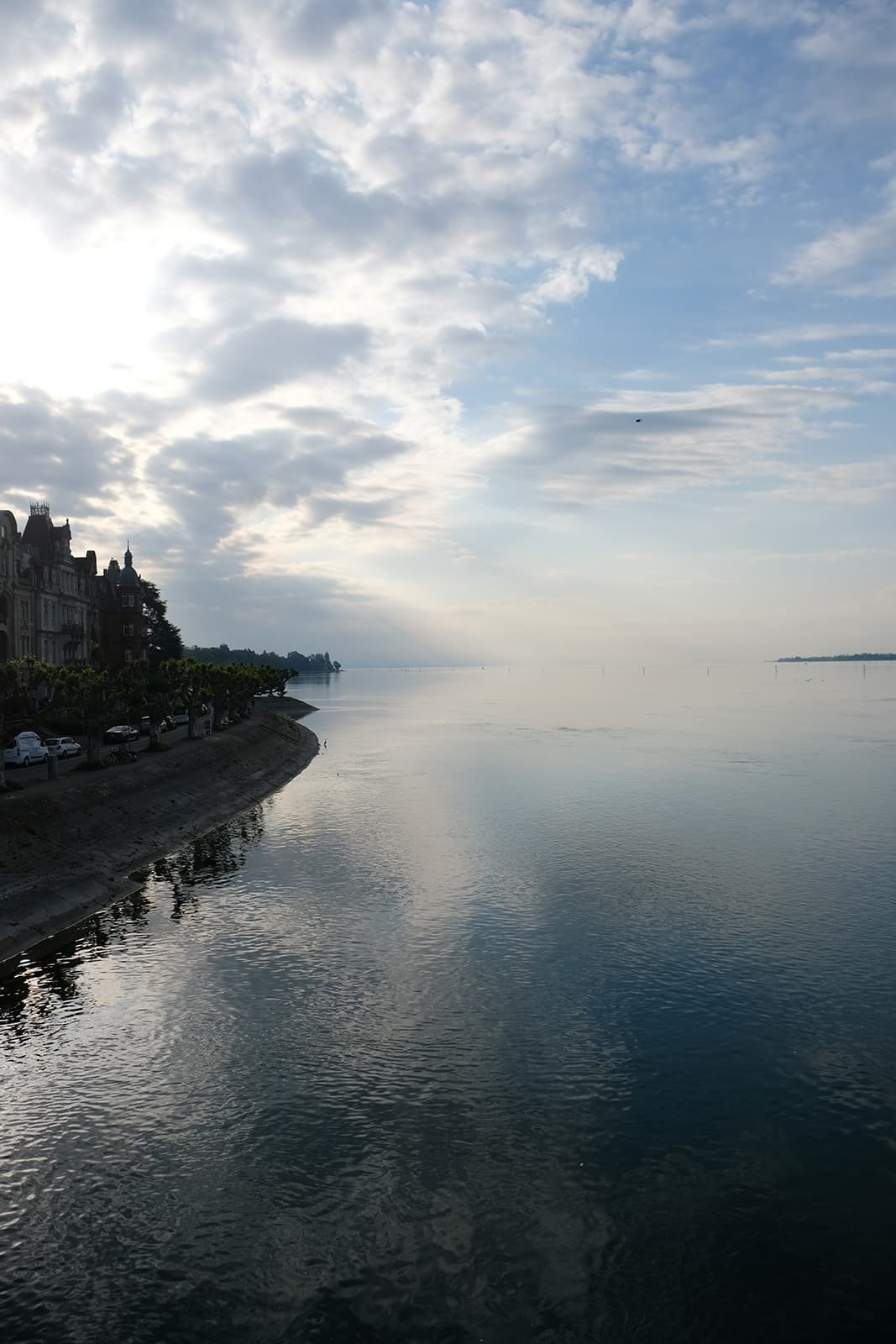 Blick auf einen Fluss bei Sonnenaufgang mit Stadtgebäuden auf der linken Seite und bewölktem Himmel.