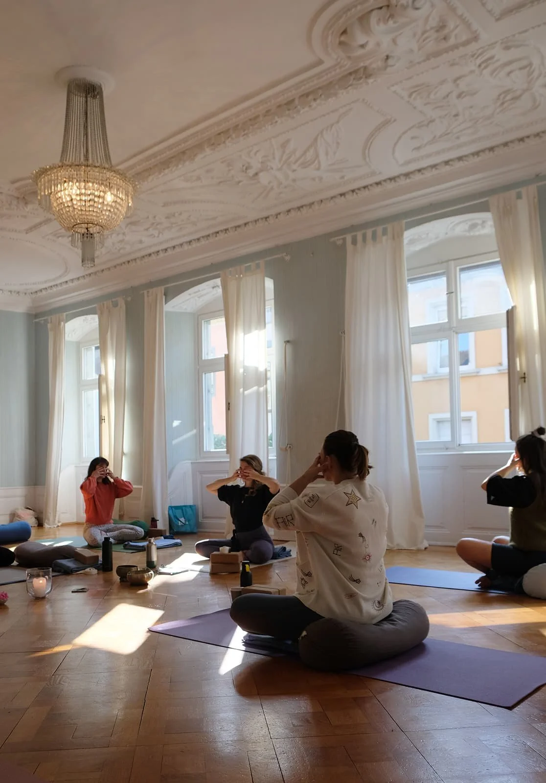 Menschen sitzen im Schneidersitz auf Yogamatten in einem hellen, elegant eingerichteteten Raum mit hohen Fenstern und einer aufwändigen Decke. Sie nehmen an einem meditativen oder Yoga-Workshop teil.
