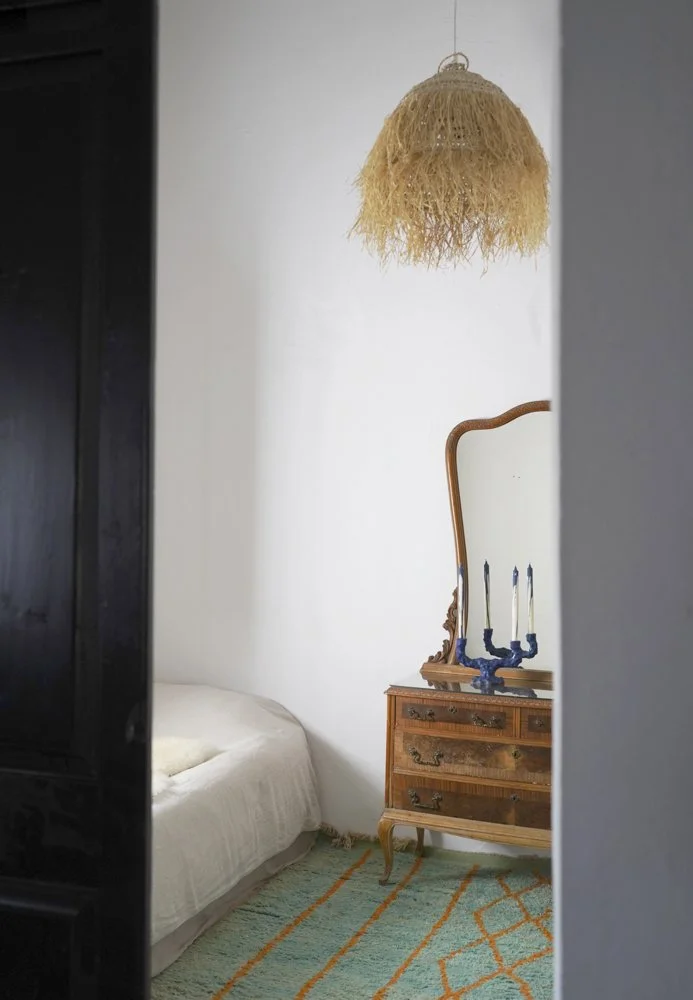 Ein Schlafzimmer mit einem weißen Bett, einem alten Nachttisch, einem ungewöhnlichen Lampenschirm aus Naturmaterialien und einem dekorativen Kerzenhalter auf dem Nachttisch.
