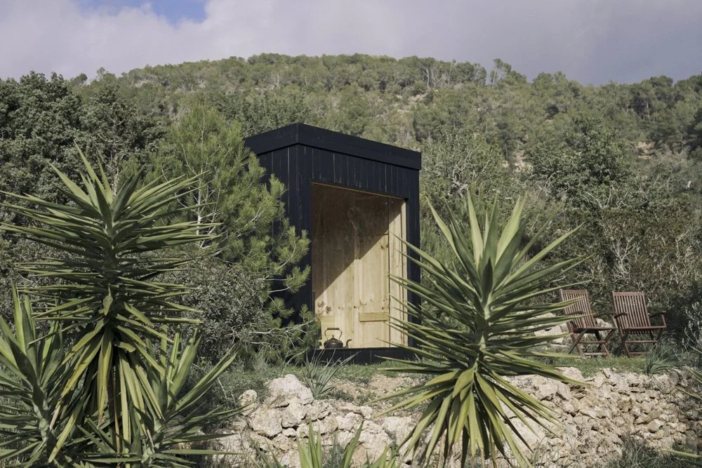 Ein kleines, schwarzes Holzhaus mit offenen Türen, umgeben von grünen Busch- und Pflanzenarten, zwei Holzstühle, im Hintergrund ein Hügel mit Bäumen, im Vordergrund.)