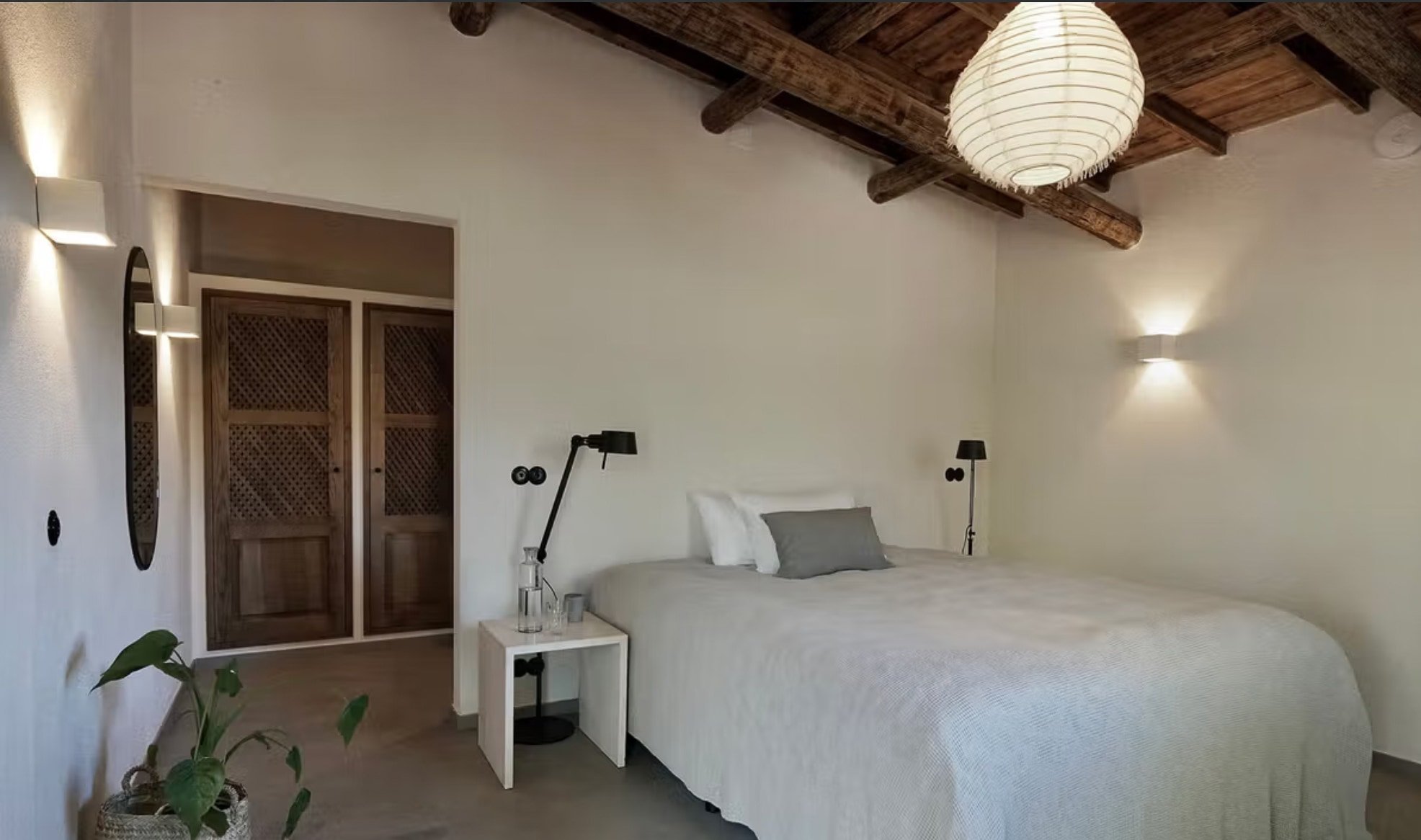 Ein Schlafzimmer mit weißem Bett, grauem Kissen, dunkler Stehlampe, Wandlampen, Holztür und einer weißen Nachttisch mit Wasserflasche, im Hintergrund sichtbares Holzdecke.