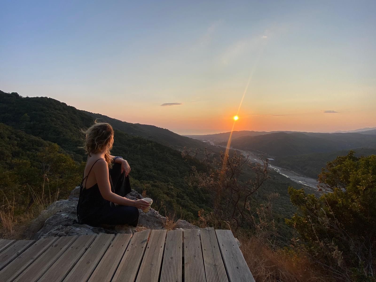 Eine Frau sitzt auf einem Felsen und blickt auf eine untergehende Sonne über einer bewaldeten Hügellandschaft und einem Fluss.