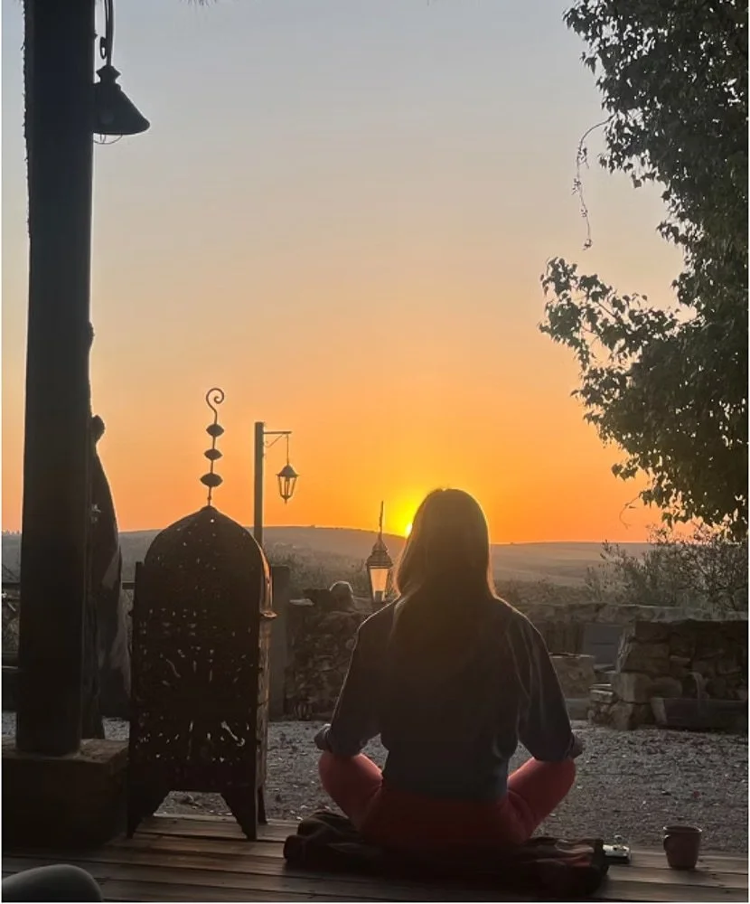 Eine Person sitzt im Schneidersitz und schaut auf einen Sonnenuntergang. Die Szene ist im Freien mit Lampen, einer Dekorationslampe und Bäumen im Hintergrund.