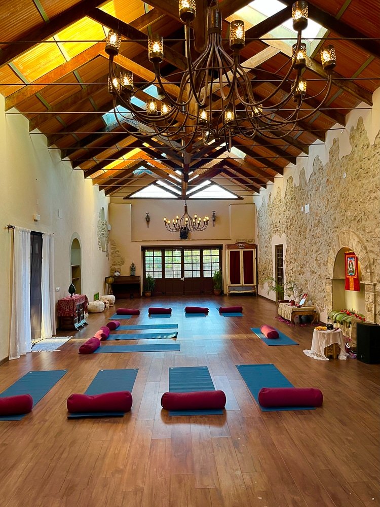 Ein großer, heller Yogaraum mit Holzfußboden, Blumenmattenträgern, und Decken und Wänden aus Stein. Der Raum ist für eine Yoga- oder Meditationstherapie vorbereitet, mit Matten und Bolstern, und hängt von zwei großen eleganten Kronleuchtern.