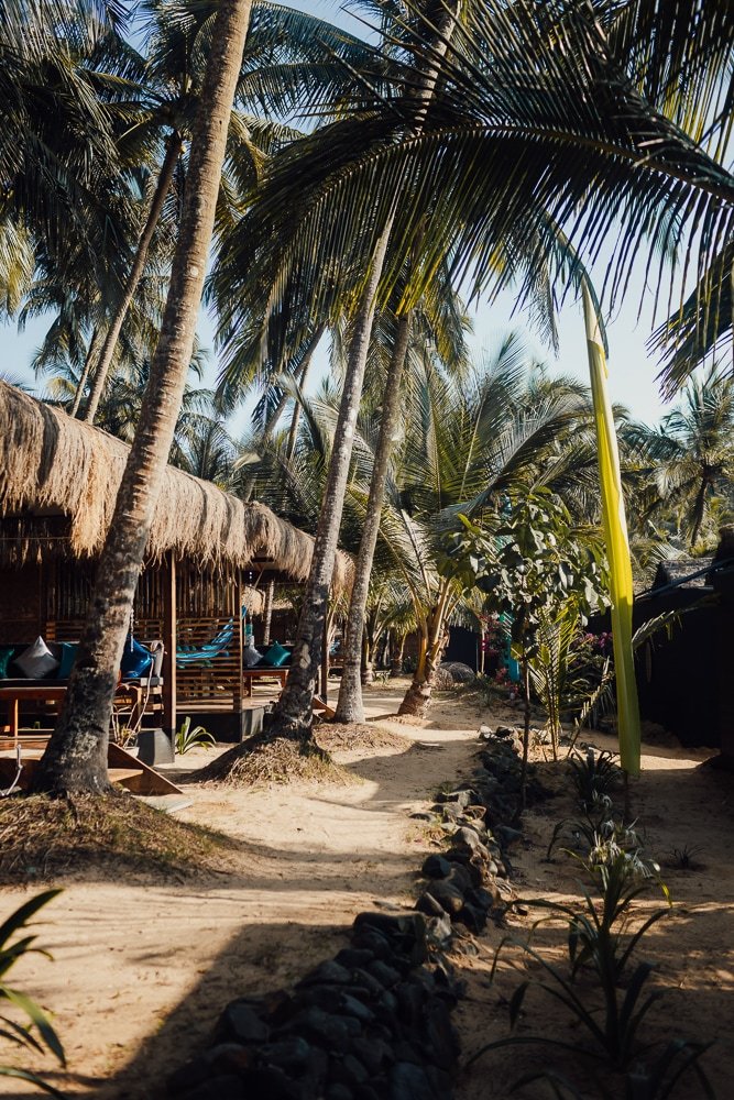 Strand mit Sandweg, palmengesäumten Hütten mit Strohdächern und Liegestühlen, tropische Pflanzen und klare Himmel im Hintergrund.