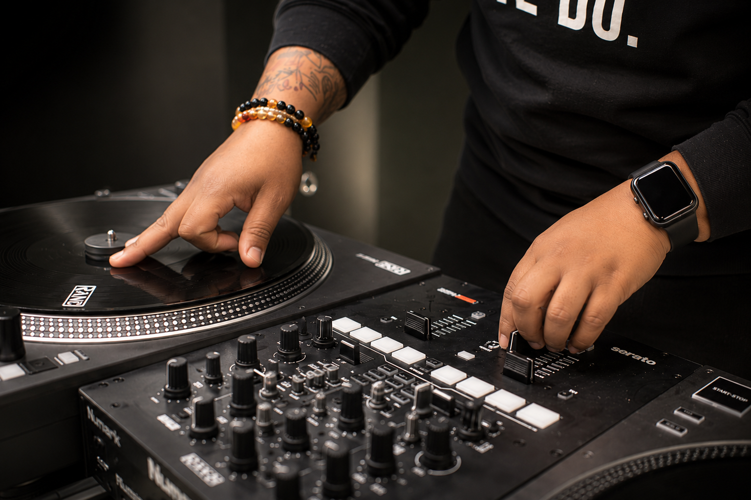 A person DJing, adjusting settings on a DJ mixer and turntable, wearing a black long-sleeve shirt and a smartwatch on the wrist.