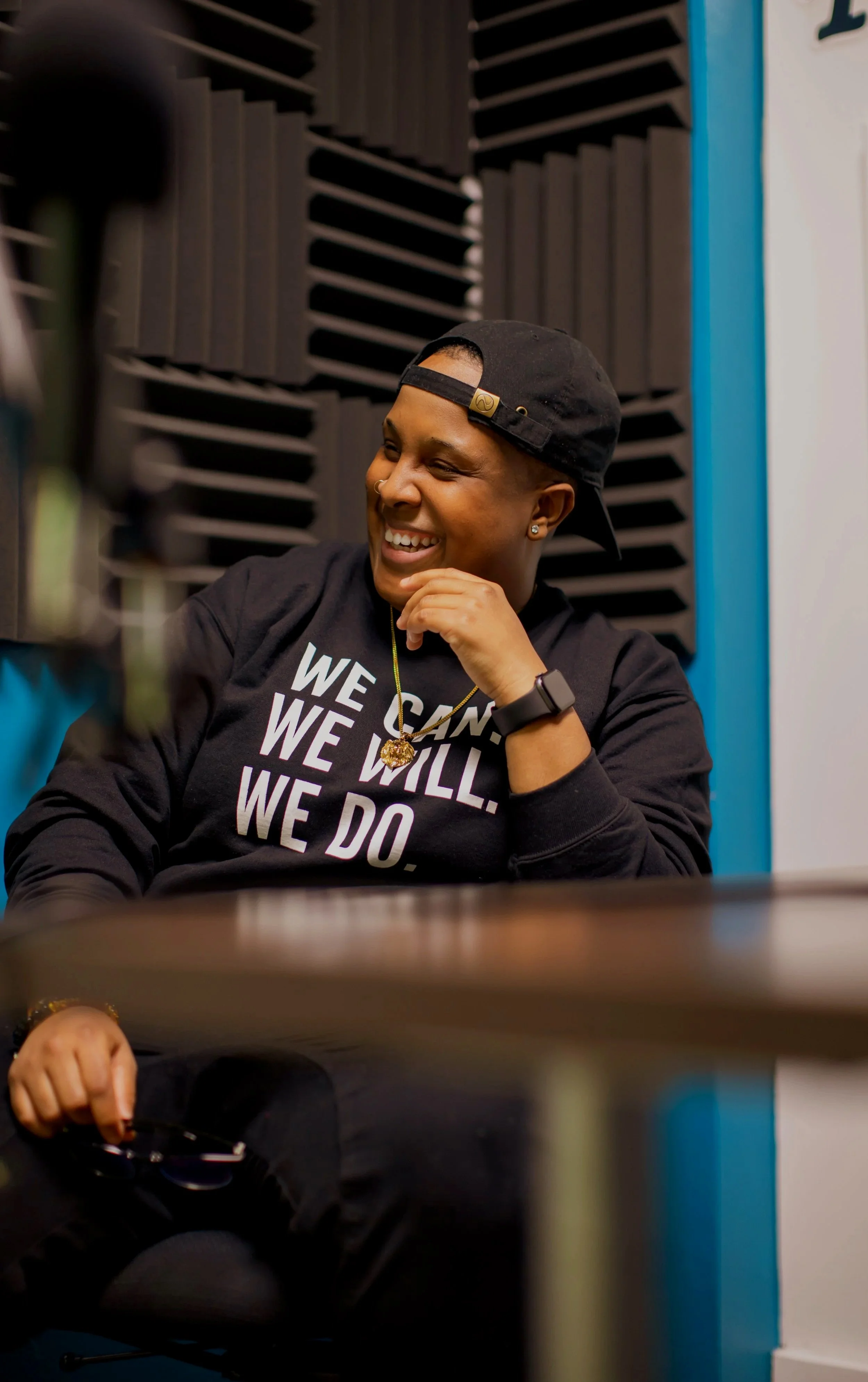 A person smiling and wearing a black baseball cap backwards, a black sweatshirt with white text, and a gold necklace, sitting in a soundproof room with black foam panels, with a table in front of them.