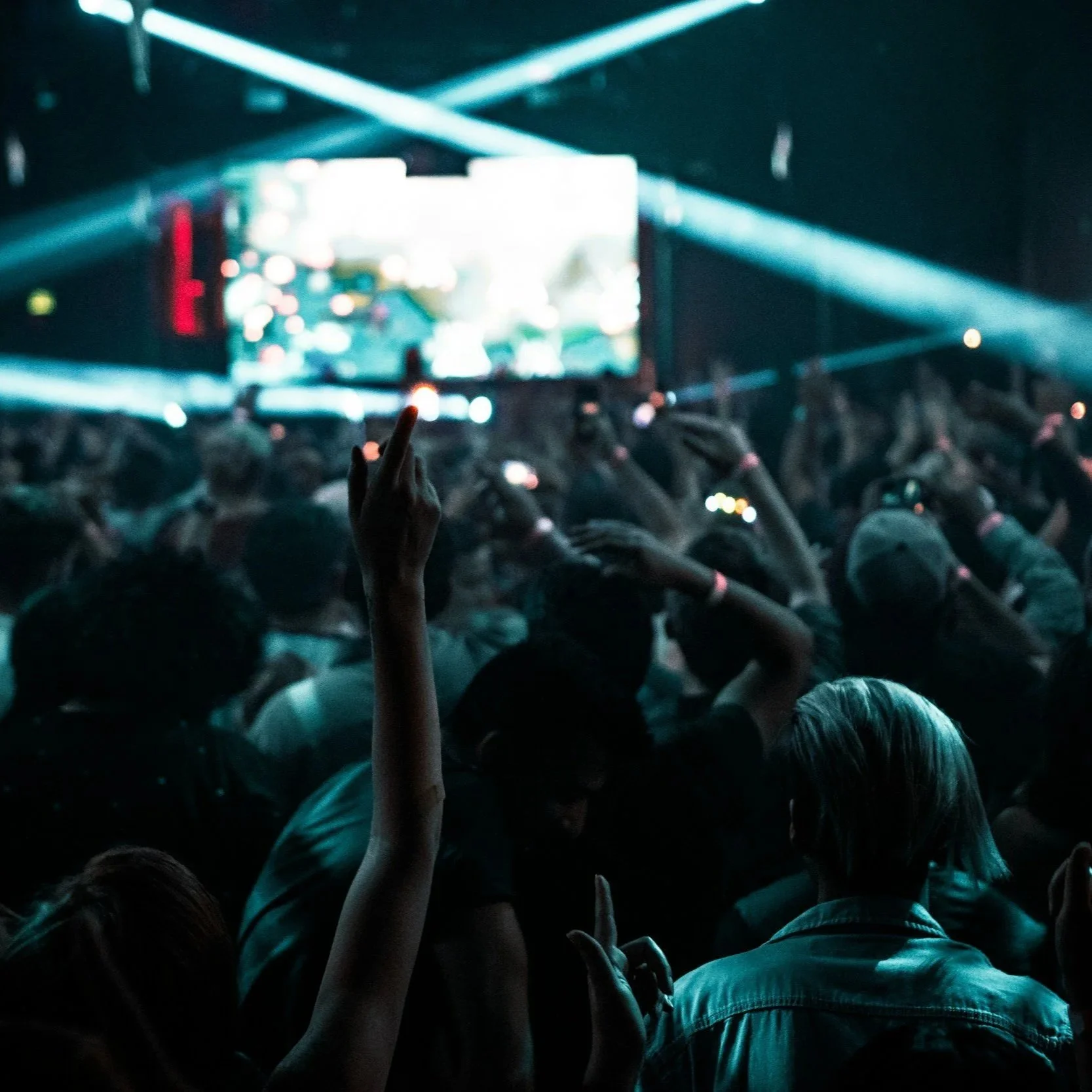 Crowd attending a concert or event in a dark venue with stage lighting and a large screen in the background, some people raising their hands.