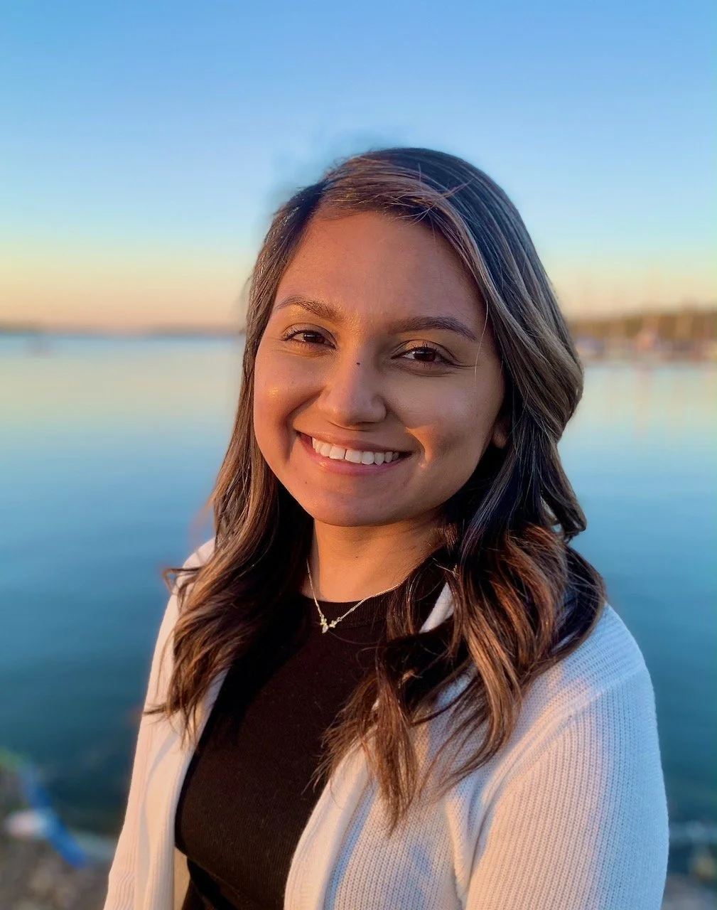 Smiling young woman outdoors near water at sunset, with colorful sky in background.