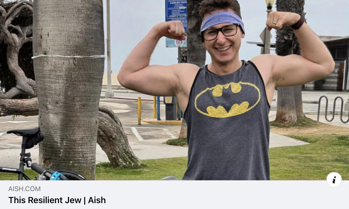 A smiling man wearing glasses, a blue headband, and a sleeveless Batman t-shirt, flexing his biceps outdoors near palm trees, with a bicycle and parking lot in the background.