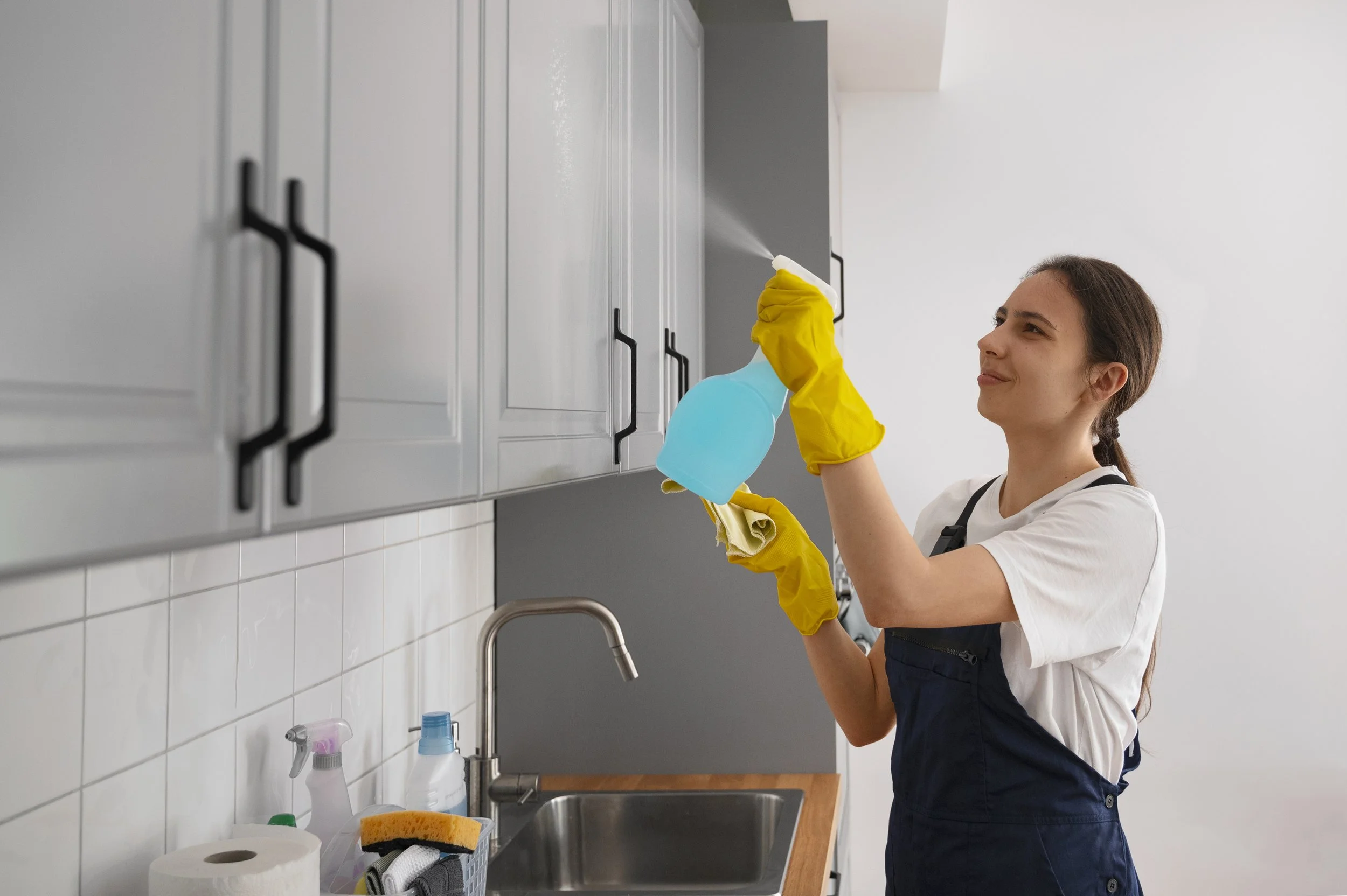 A woman cleaning kitchen cabinets with a spray bottle and cloth, wearing yellow rubber gloves.