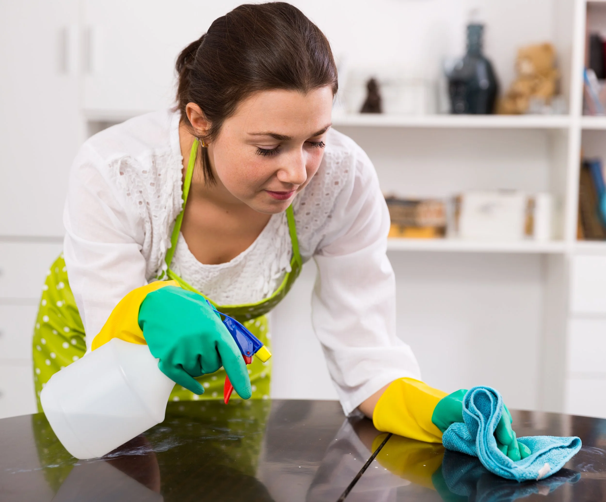 A woman in a white top, yellow rubber gloves, and an apron cleaning a dark countertop with a cloth, in a kitchen setting with shelves and decor in the background.