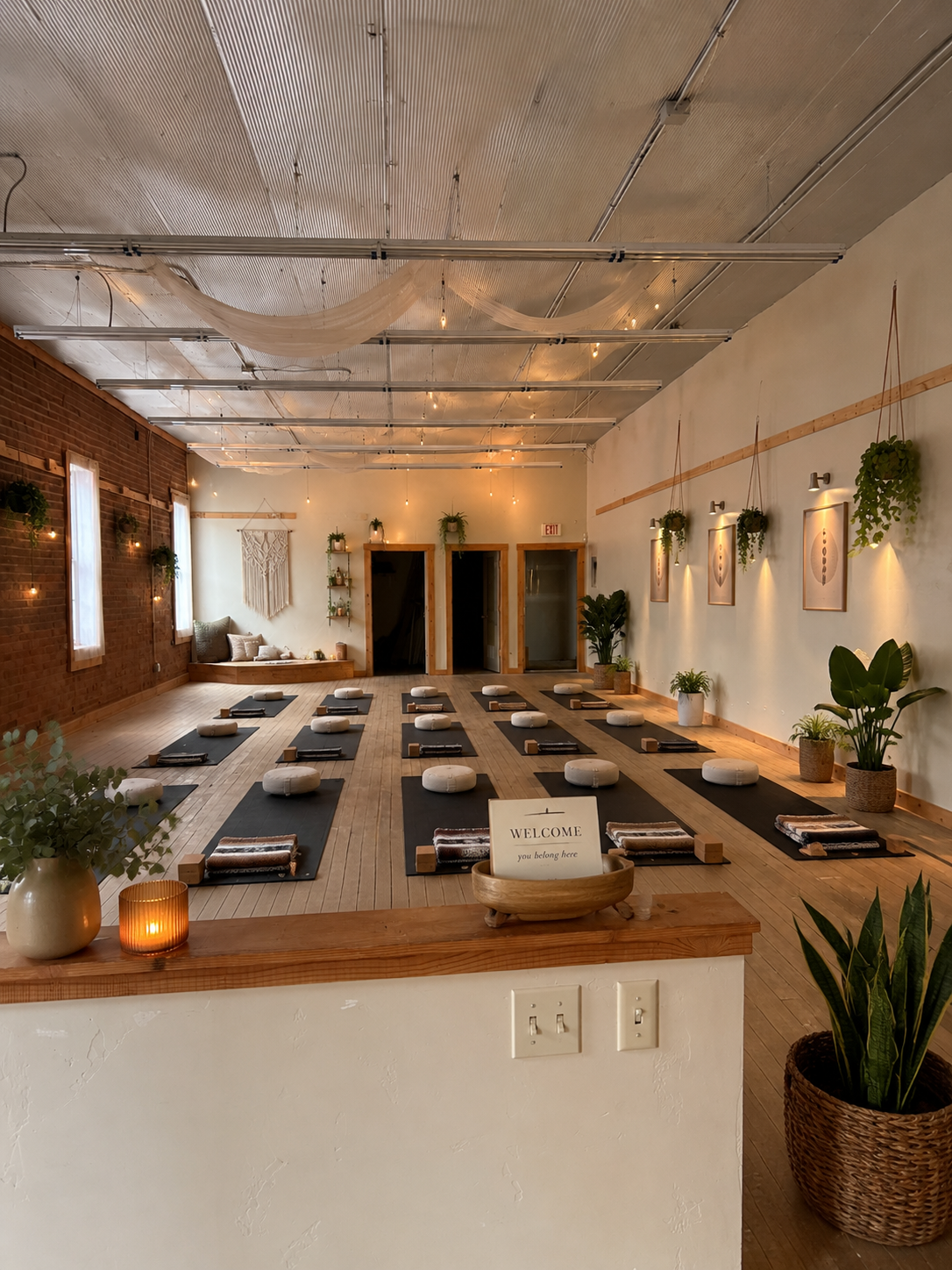 A yoga studio with black mats, white cushions, and blankets arranged on a wooden floor, decorated with plants, framed artwork, and soft lighting.