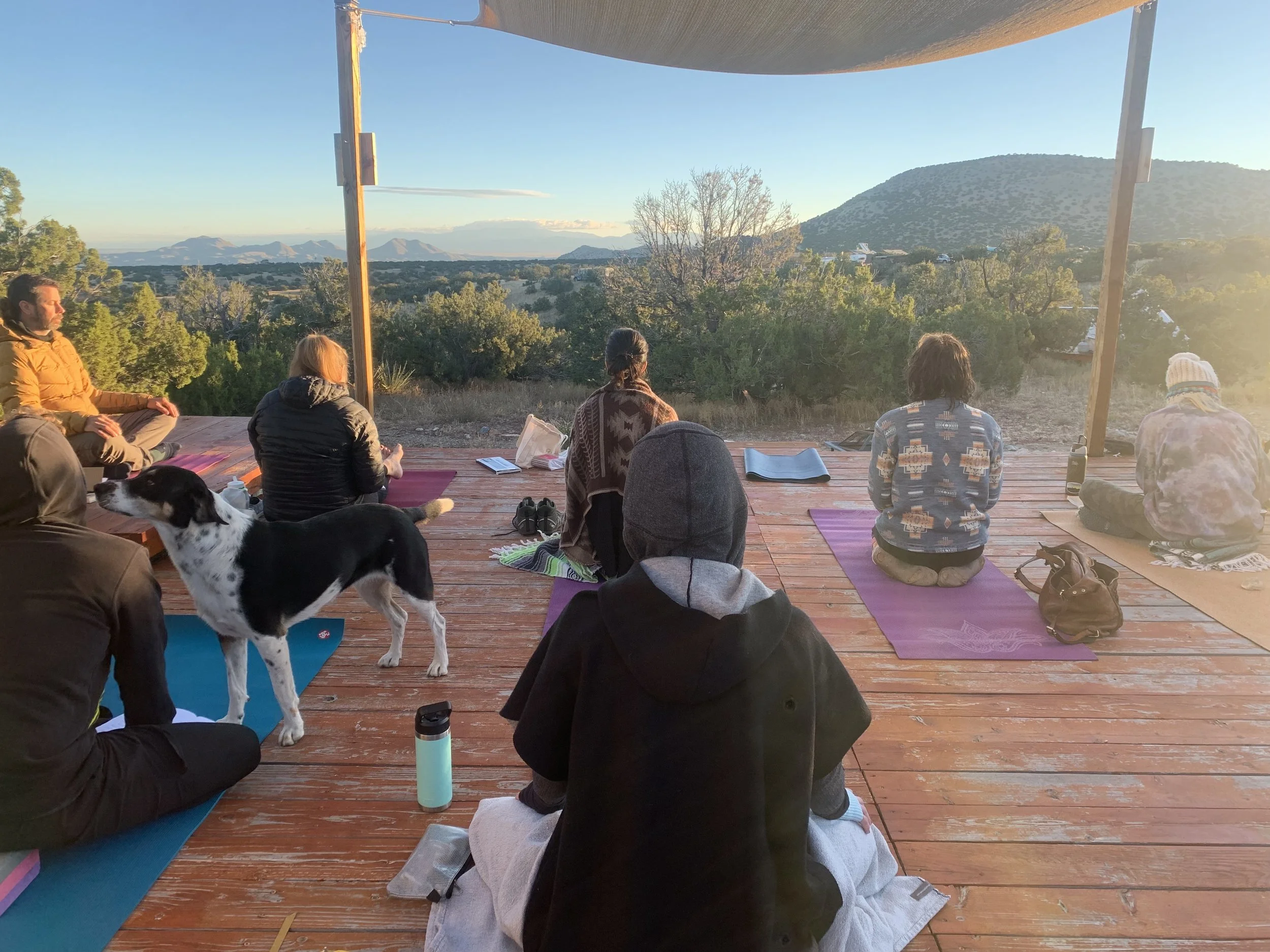 People practicing yoga outdoors on a wooden deck with a scenic mountain view during sunset.