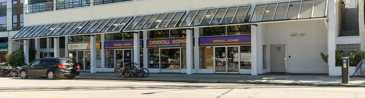 Street view of a game store named DREXOLL GAMES with large windows, purple and yellow signage, and bicycles parked outside. There are cars parked along the street and a parking meter on the sidewalk.