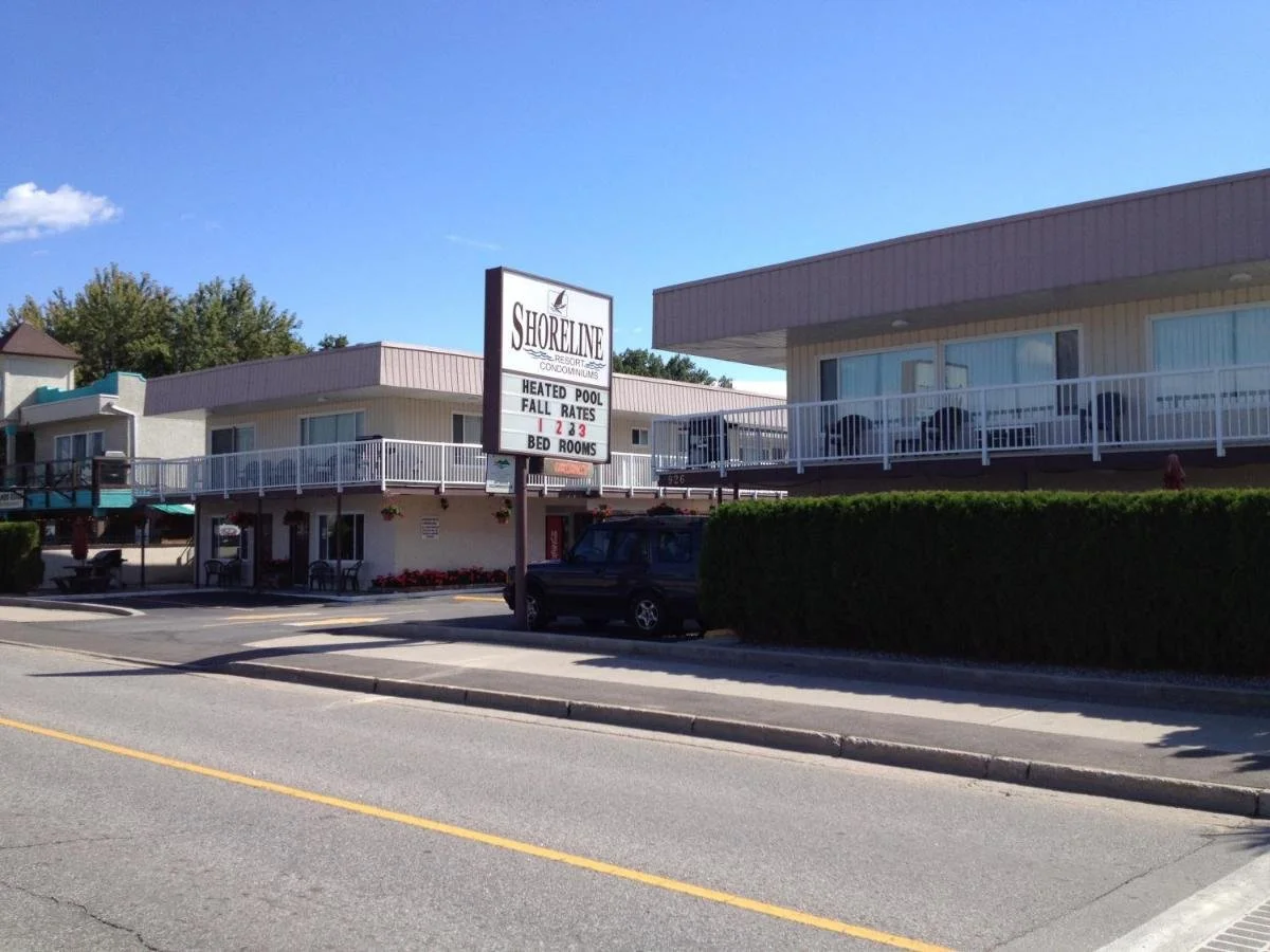 Exterior view of Shoreline Resort with balconies, parking lot, sign advertising heated pool, fall rates, and bed rooms, a black vehicle parked in front, clear blue sky, and trees in the background.