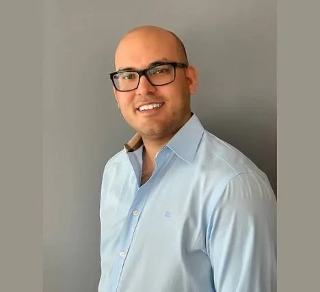 A man with glasses, a bald head, and a light blue dress shirt smiling for the photo, standing against a plain gray background.