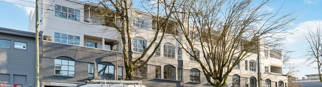 A street view of a multi-story building with commercial space on the ground floor, with leafless trees in front and a partly cloudy sky overhead.