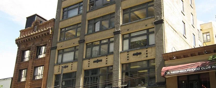 Tall urban building with multiple floors, windows, and worn exterior, adjacent to historic brick building with decorative architectural details, and a street scene with power lines and a neighborhood club sign.
