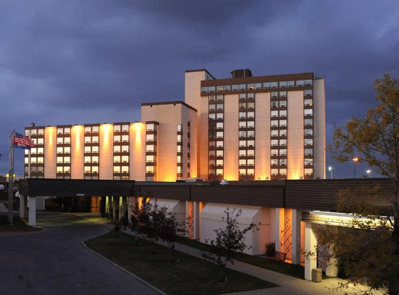 A multi-story hotel building illuminated at night with yellow lighting, with an American flag on a flagpole in front and dark cloudy sky above.