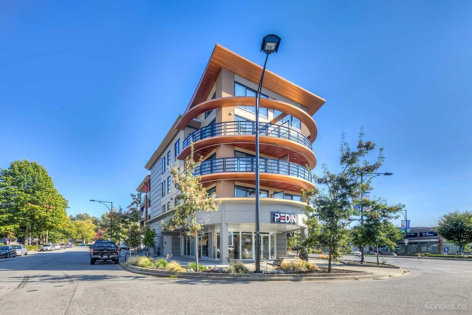 A modern four-story commercial building with curved balconies, glass railings, and a sign that reads "PEDINI" at the ground level, surrounded by trees, parked cars, and streetlights against a clear blue sky.