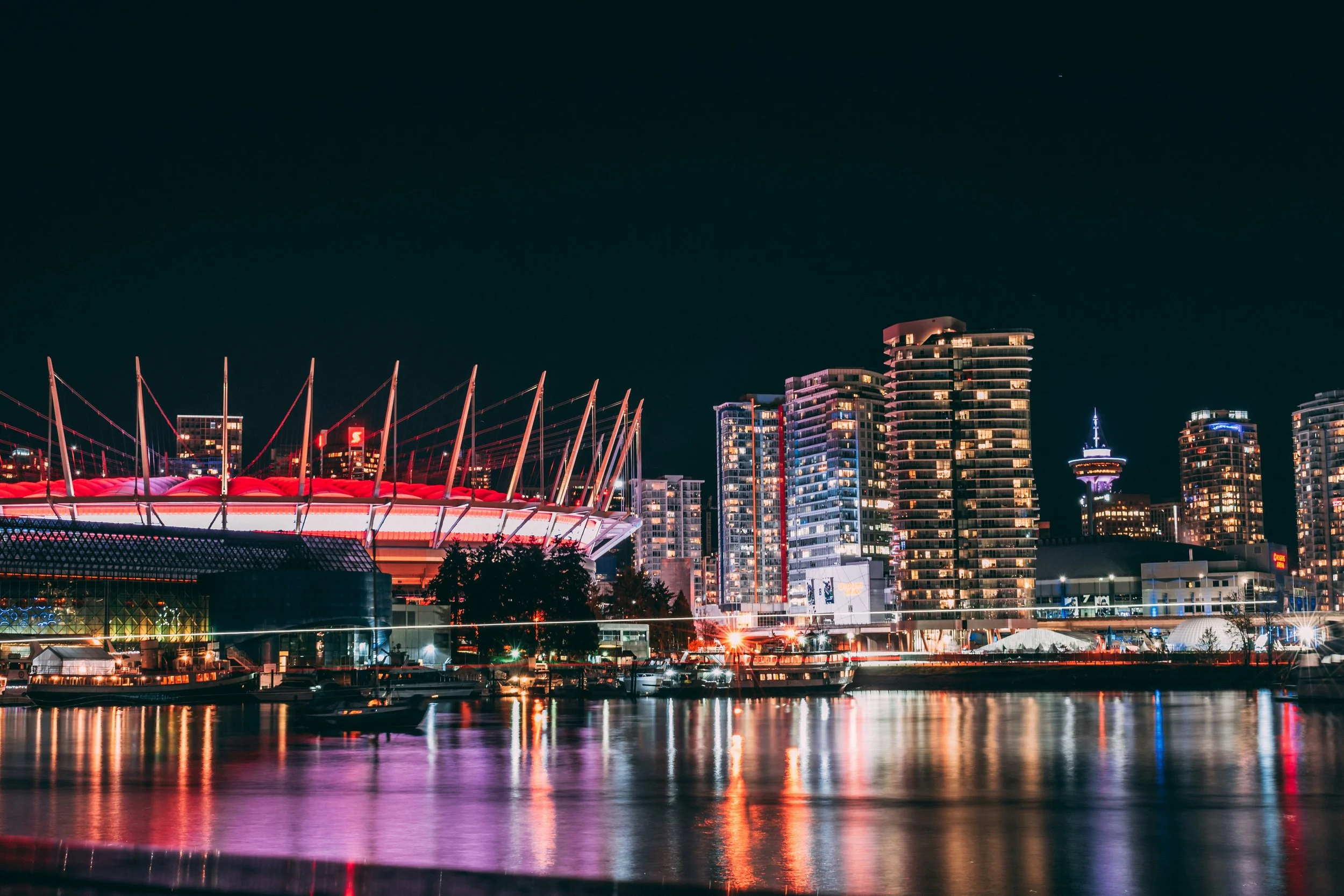 Nighttime view of a city skyline with illuminated modern buildings and a brightly lit stadium reflecting on the water.